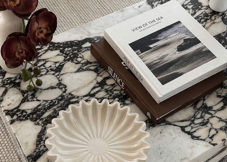 Decorative bowl on a marble surface with a book titled 'View of the Sea' and flowers.